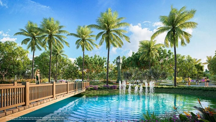 Cầu gỗ bắc qua hồ nước có đài phun nước, hàng dừa và cây xanh trong khuôn viên dự án